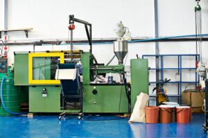 Industrial laboratory equipment manufacturing setup featuring an injection molding machine with a green and yellow frame. The machine includes a metal hopper for feeding raw materials and a conveyor belt for finished parts. The workspace has blue flooring, organized storage shelves, and multiple containers of raw materials, emphasizing efficient lab equipment production processes.